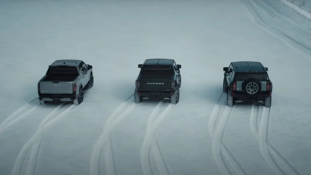 Three GMC Hummer EVs demonstrating King Crab mode by driving diagonally across a snowy field, leaving distinct tire tracks.